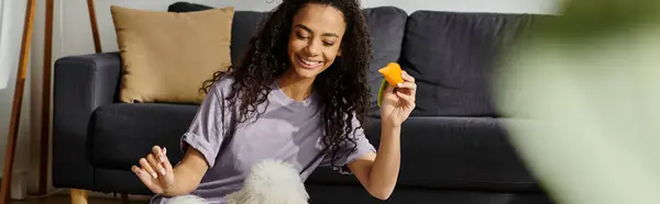 stock image A young woman relaxes on a couch, holding a piece of toy fruit while her bichon frise dog sits beside her.