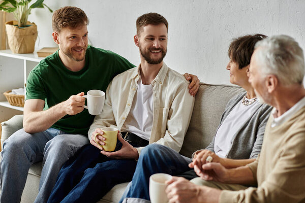 A gay couple sits with parents on a couch in a living room, enjoying a conversation and cups of coffee.