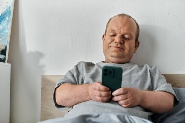 Kapsayıcı bir adam yatakta gevşer, akıllı bir telefon tutar ve gülümser, güne başlar..
