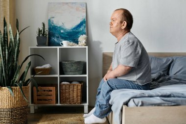 Kapsayıcılığı olan bir adam yatağın kenarında oturur, içe dönük görünür..