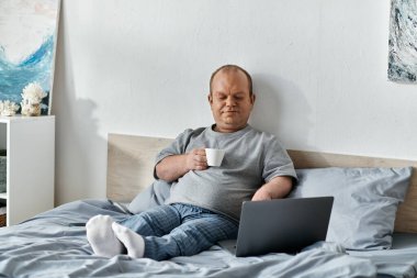Kapsayıcı bir adam yatakta oturur, laptopunu kullanırken bir fincan kahvenin tadını çıkarır..