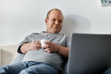 Kapsayıcı bir adam bir yatakta oturuyor, elinde bir kahve fincanı ve dizüstü bilgisayara bakıyor..