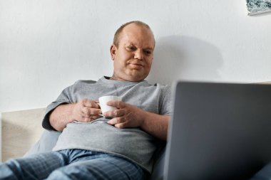 A man with inclusivity is sitting on a bed, holding a coffee cup and looking at a laptop. clipart