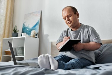 A man with inclusivity sits on his bed, writing in a notebook, with a laptop beside him. clipart