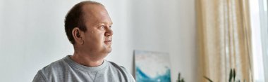 A man with inclusivity in a grey shirt sits indoors, looking thoughtfully out of the window. clipart