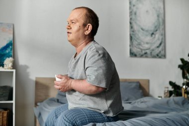 Kapsamlı bir adam yatak odasındaki bir yatağa oturur, elinde bir kupa tutar ve içe dönük görünür..