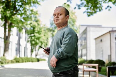 Kapsamlı yeşil kazak giyen bir adam elinde telefonuyla şehir caddesinde yürüyor..