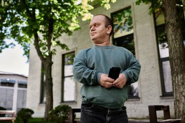 Kapsamlı bir adam bir binanın önünde duruyor, telefonu tutarken düşünceli görünüyor..