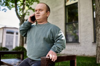 Kapsamlı bir adam bankta oturuyor, telefonuyla konuşuyor, sakin bir açık hava ortamının ortasında..