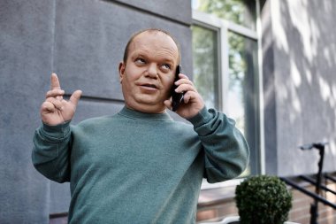 Kapsayıcı bir adam yeşil bir sweatshirt giyerek sokakta dikiliyor, telefonla konuşuyor, muhtemelen bir noktaya parmak basıyor ya da sözlerini vurguluyor..