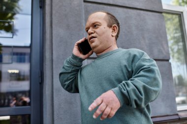 Yeşil süveter giyen bir adam telefonunu binanın dışında kullanır..