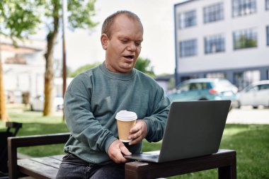 Kapsayıcılığı olan bir adam parkta bir bankta oturur, dizüstü bilgisayarıyla çalışır ve kahvenin tadını çıkarır..