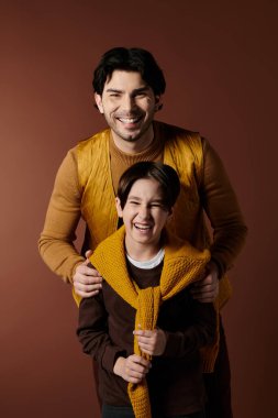 A father and son stand close together, both smiling and laughing. The son is holding a yellow scarf, and the father has his arm around the sons shoulders. clipart