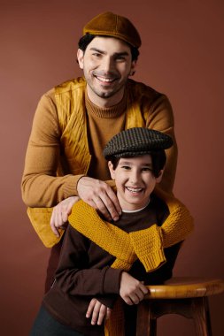Bir baba ve oğul birbirlerine yakın dururlar, içten gülümserler, kahverengi ve sarı elbiseler giyerler..