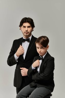 A father and son pose in matching tuxedos, the son sitting, the father standing. clipart