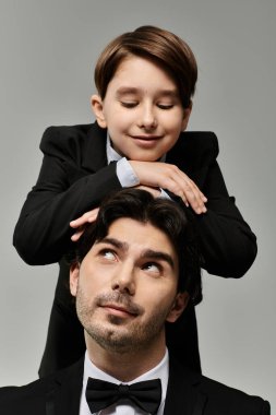Genç bir çocuk ellerini şakacı bir şekilde babasının kafasına koyar, baba ise gülümseyerek yukarı bakar..