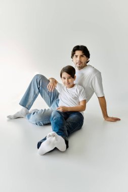 Bir baba ve oğlu beyaz gömlek ve kot pantolon içinde beyaz bir zeminde birlikte oturuyorlar..