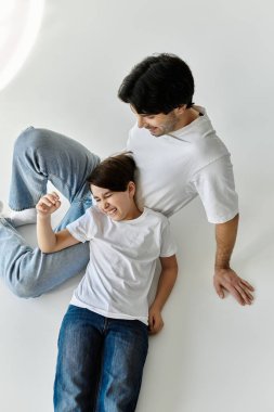 Bir baba ve oğul bir an için neşe ve kahkahayı paylaşıyor, beyaz bir zeminde yatıyor..