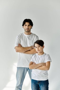 Bir baba ve oğlu, beyaz tişört ve kot pantolon giyip kollarını kavuşturarak yan yana duruyorlar..