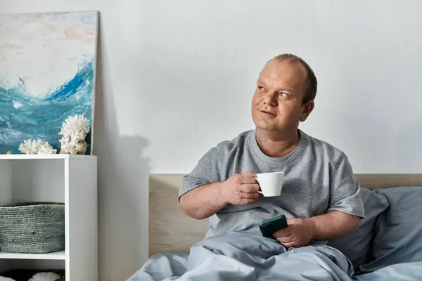 stock image A man with inclusivity sits in bed, enjoying a cup of coffee and looking thoughtful.