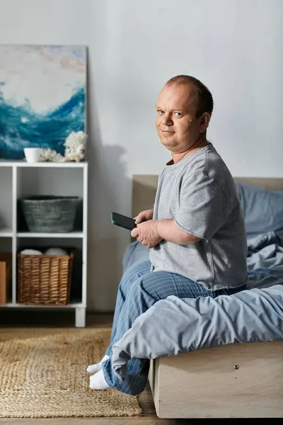 stock image A man with inclusivity sits on his bed, looking at his phone. He is wearing casual clothing and seems relaxed.