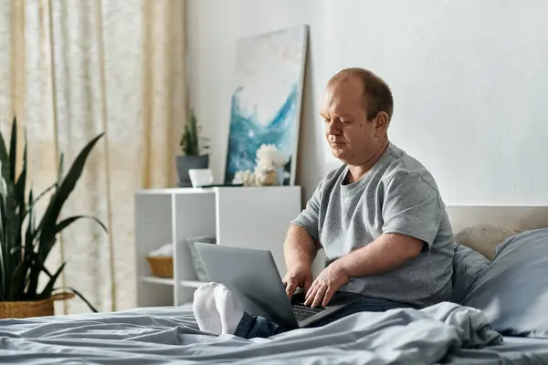 stock image A man with inclusivity sits on his bed in a bright room, working on his laptop.