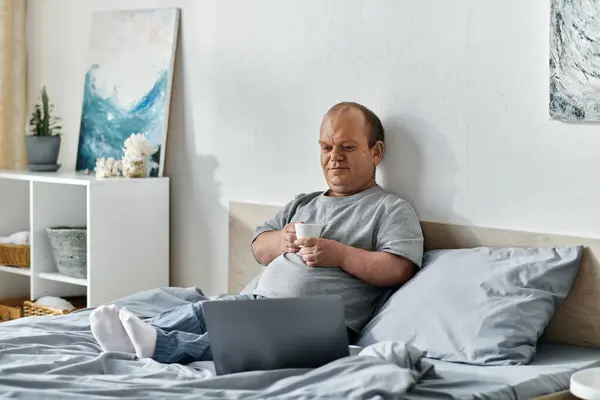 stock image A man with inclusivity sits in bed, holding a cup of coffee and looking at his laptop.