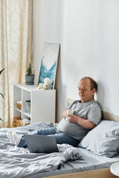 A man with inclusivity sits in bed, holding a cup of coffee, and using a laptop.