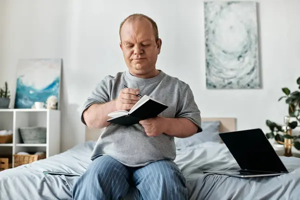 stock image A man with inclusivity sits on a bed writing in a notebook, with a laptop nearby.