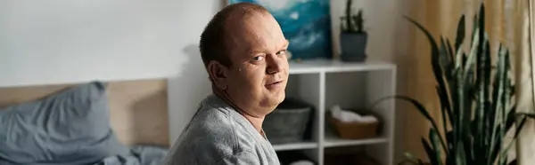 stock image A man with inclusivity sits on a bed in a well-lit room, looking towards the camera with a thoughtful expression.