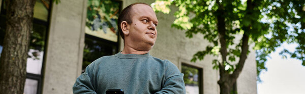 A man with inclusivity in a blue sweater stands in front of a building, looking thoughtfully towards the street.