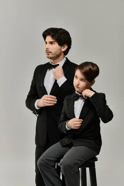 stock image A father and his son stand side by side, both dressed in black suits and bow ties, ready for a formal event.
