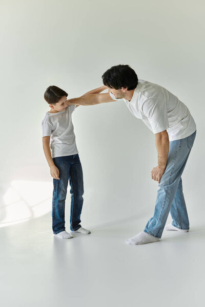 A father and son stand in a white room, the father leaning down towards the boy, suggesting a moment of playful interaction.