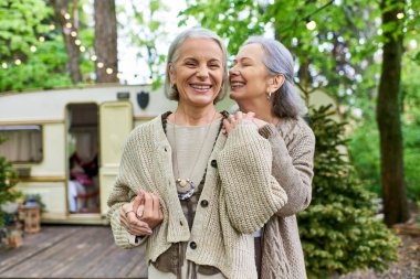 Two middle-aged women, a lesbian couple, share a laugh while camping in a lush green forest. A camper van is visible in the background. clipart