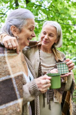 İki orta yaşlı kadın kamp gezisinde sıcak bir içecek içerken birlikte gülüyorlar..