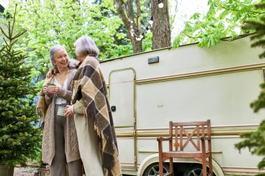 Orta yaşlı lezbiyen bir çift sıcak bir içki içip, karavanlarının önünde, yemyeşil bir ormana kurulmuş bir şekilde vakit geçiriyorlar..