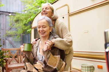 A lesbian couple laughs together while enjoying a cup of coffee outside their camper van on a sunny day. clipart