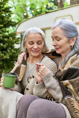 A lesbian couple enjoys a cozy moment together while camping in a lush green forest. They are wrapped in blankets and share a warm drink. clipart