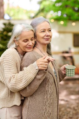 Two middle-aged women embrace in a forest campsite, near their van, showcasing love and affection in a natural setting. clipart