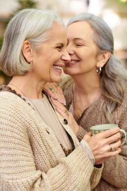 Two women, an older lesbian couple, are close together in a forest setting. They appear to be camping, and one of them is holding a mug. clipart