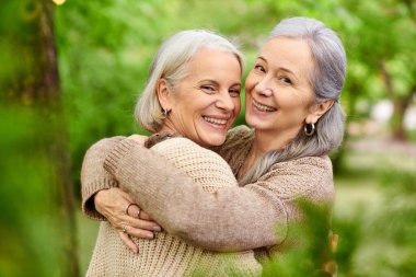 Two middle-aged women embrace in a green forest setting. They appear happy and relaxed, enjoying their time outdoors. clipart