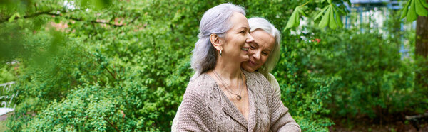 Two middle-aged women in cardigans embrace each other in a verdant setting.