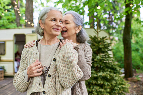 Two middle-aged women share a loving moment in a forest setting, near a camping van.