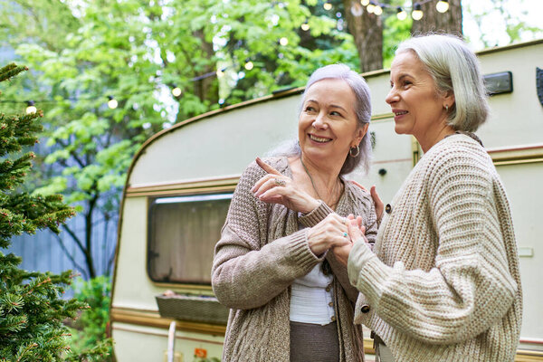 A lesbian couple smiles and holds hands in a verdant forest near a camping van.
