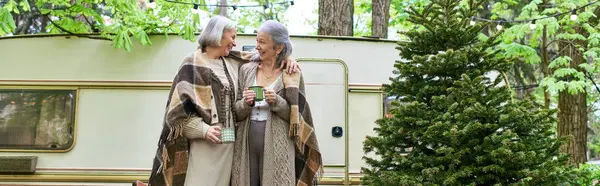 stock image Two middle-aged women wrapped in blankets, enjoy a cup of coffee while camping near their van in a lush forest.