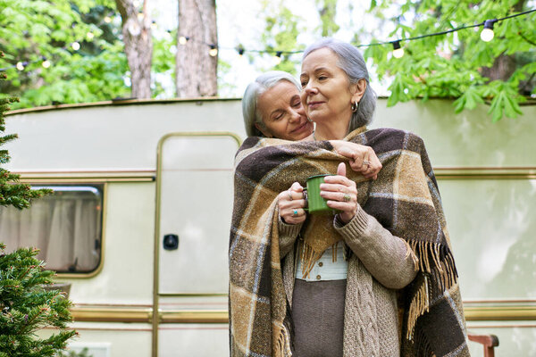 Лесбийская пара, завернутая в одеяло, наслаждается теплым напитком во время лагеря в зеленом лесу возле каравана.