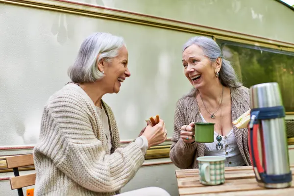 stock image Two women, a middle-aged lesbian couple, laugh together while enjoying a snack and drinks outside their van on a camping trip.