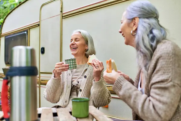 stock image Two middle-aged women enjoy a coffee break while camping in the forest, laughing and sharing a meal.