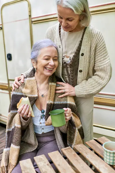 Stock image A lesbian couple enjoys a cozy moment together while camping in a lush green forest, sharing a meal and a drink.