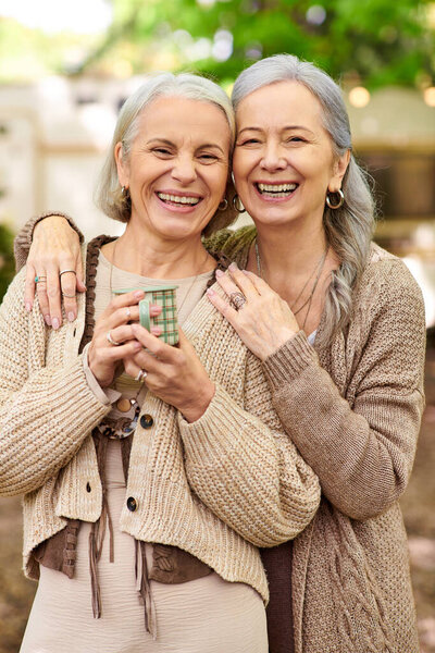 Две улыбающиеся женщины средних лет изображены обнимающимися в зеленом лесу. Они разбивают лагерь вместе, на заднем плане виден фургон.